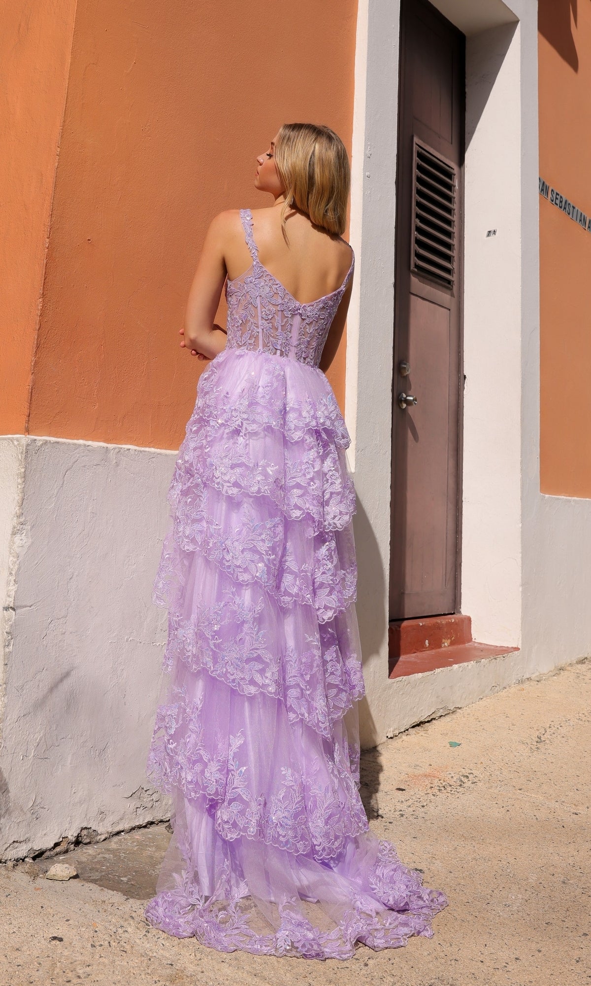 A woman in the Long Lace Ruffled A-Line Prom Dress T1335, featuring layered lavender lace, stands with her back to the camera, facing a tan and white building with a brown door on a sunny day.